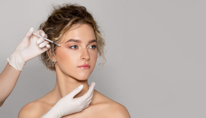 Young beautiful woman getting botox injection for eyes area, standing over grey studio background,...