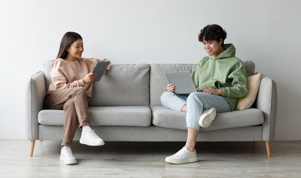 Handsome young Asian guy and his girlfriend using modern gadgets while sitting on couch at home, copy space. Full length of millennial couple communicating online via laptop and tablet pc