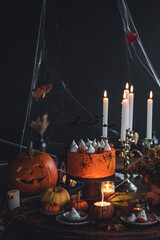 Beautiful table with homemade delicious cake for Halloween party with meringue ghosts and chocolate pumpkins. Candles in chandelier, spider web, jack-o-lantern for festive holiday decoration. Dark 