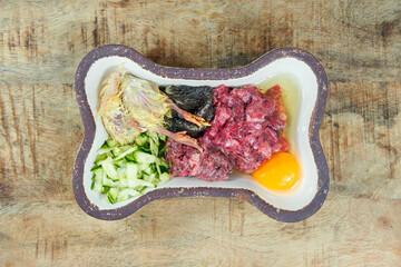 Top view of a natural raw dog food in a bone-shaped ceramic bowl on a rustic wooden surface. Raw minced meat, egg, whole unplucked day-old chick, tripe.