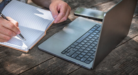 Businessman or freelancer writing in notebook while working outdoors on laptop at wooden table. Creative remote work or planning concept