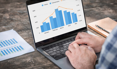 Male entrepreneur analyzing monthly conversion and click-through analytics on laptop at wooden table workspace. Remote work and data analysis concept