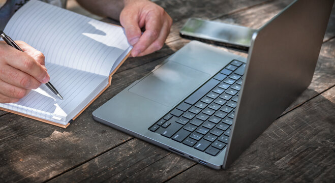 Businessman or freelancer writing in notebook while working outdoors on laptop at wooden table. Creative remote work or planning concept