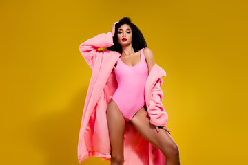 Confident young woman wearing pink bodysuit and coat, posing gracefully against a vibrant yellow...