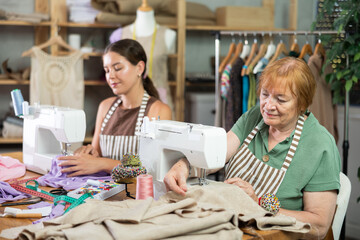 Two seamstresses work together and sew a collection of clothes on sewing machines in the workshop. Female dressmakers sitting at a table and sewing clothes to order