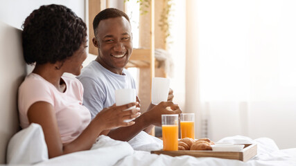 Honeymoon concept. Happy black young couple sitting in bed in morning and having breakfast, side...