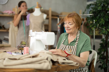Mature dressmaker in an apron sits at a table and sews clothes against the background of an assistant woman who works with a mannequin. Dressmakers sew custom-made clothes