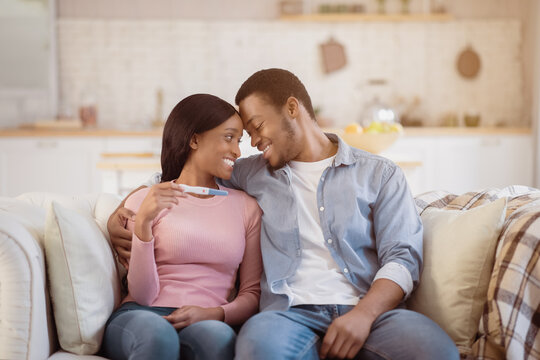 Loving African American couple with positive pregnancy test cuddling on sofa at home. Millennial black man with his wife feeling happy to have baby, having romantic moment together
