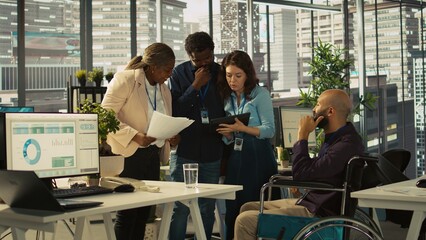 Employees and coworker with disability looking over revenue figures in inclusive office. Man in wheelchair working together with colleagues, comparing company statistical numbers, camera B