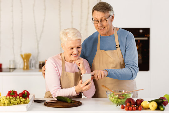 Cheerful Senior Spouses Using Cellphone Cooking Dinner Together And Browsing Internet For Online Recipes Standing In Modern Kitchen At Home. Elderly Couple Using Smartphone Preparing Meal Concept