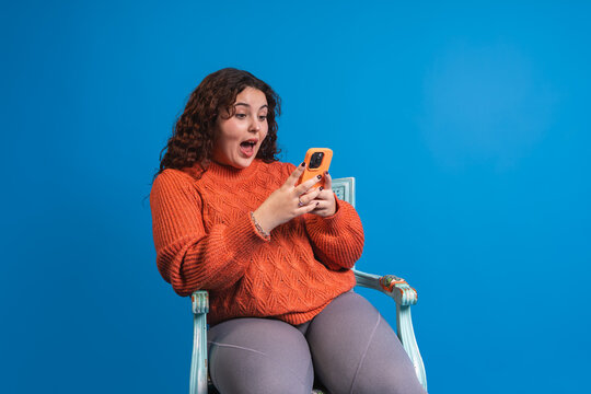 Shocked plus size woman using mobile phone, surprised by unexpected news on blue background - Powered by Adobe