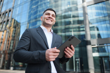 Fototapeta premium Professional Young Man Smiles While Using a Tablet Outside a Modern Skyscraper on a Sunny Day