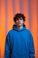 Obraz premium A portrait photograph of a young man wearing a bright blue hooded sweatshirt against a vibrant orange and pink gradient background.