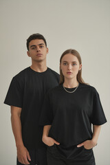 A minimalist studio portrait of two young people standing against a plain off-white wall. Both are wearing plain black t-shirts.