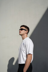 A minimalist portrait photograph of a young man in profile view, looking upward against a plain light gray wall. He wears a plain white crew neck t-shirt and black sunglasses. 