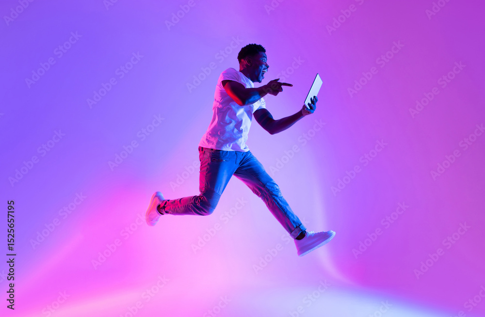 Aufkleber excited young black guy with tablet computer jumping in neon light, copy space. emotional african am