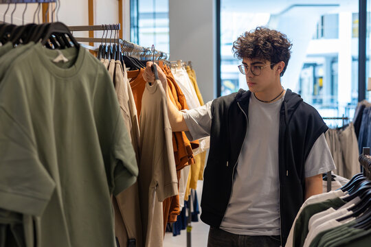 Teen boy choosing stylish t-shirt in clothing store. Young customer shopping trendy casual wear at fashion retail shop. Boy shopping alone in urban fashion shop. Copyspace - Powered by Adobe