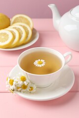 Delicious chamomile tea in cup, fresh flowers and lemon on pink wooden table, closeup