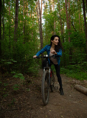 Naklejka premium Beauty smiling woman biking on bicycle on spring trees green forest background, looking happy. Enjoying fun girl outdoors. Female cyclist riding in park in sport fashion clothes. Full lenght portrait
