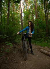 Naklejka premium Beauty smiling woman biking on bicycle on spring trees green forest background, looking happy. Enjoying fun girl outdoors. Female cyclist riding in park in sport fashion clothes. Full lenght portrait