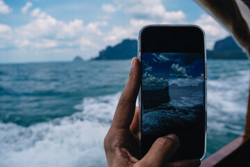 Hand of tourist holding smartphone taking picture of seascape with mountains and cloudy sky from boat