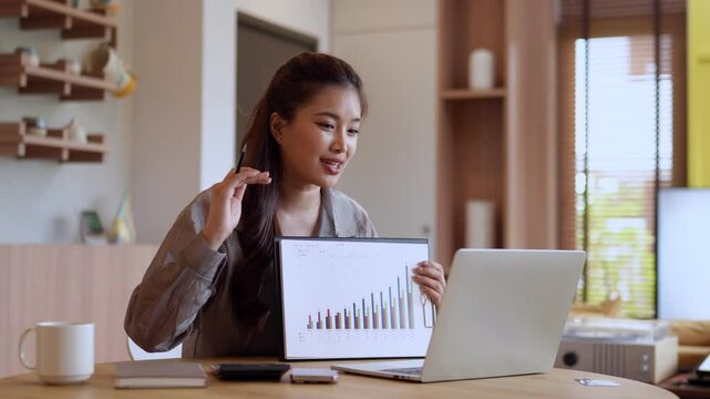 Asian businesswoman confidently presenting data chart through online video call from home office, combining professional and modern digital lifestyle.