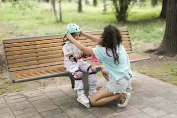 Young mother putting a helmet on her daughter before rollerblading. Safety first! A loving and caring moment on a bench in a green park.