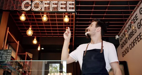 Positive young bearded male barista in white t shirt apron and reusable face mask smiling and looking at camera while standing and point to the inscription coffee in modern coffee house. - Powered by Adobe