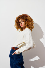 Fashionable young woman with curly hair wearing a white sweater against a plain background,...