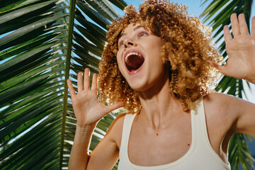 Happy woman with curly hair enjoying sunny tropical environment, surrounded by green palm leaves...