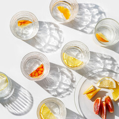 Citrus Infused Water in clear Glasses and plate,  artistic light reflections and shadow in natural sunlight. Summer drink concept, minimal pattern, flavored water with yellow red slices of fruit