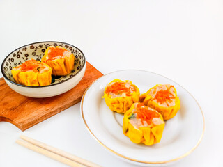 Yellow dimsum or Siomay chicken and shrimp variance with carrot above served on white plate,chopsticks, wooden cutting board, in white background
