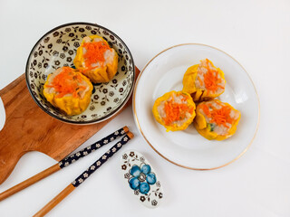 Yellow dimsum or Siomay chicken and shrimp variance with carrot above served on white plate,chopsticks, wooden cutting board, in white background
