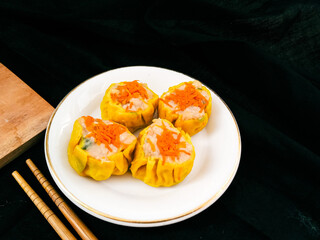 Yellow dimsum or Siomay chicken and shrimp variance with carrot above served on white plate,chopsticks, wooden cutting board, in black background
