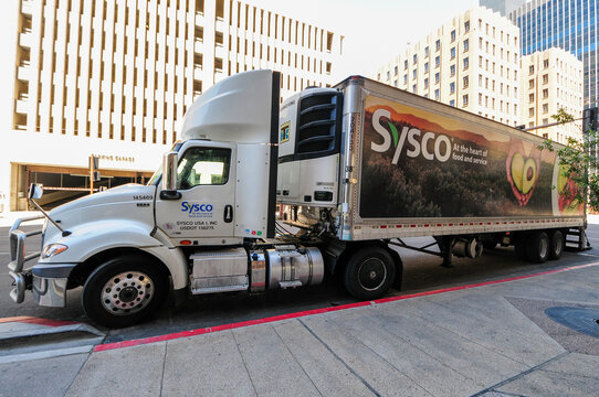 Sysco refrigerated food delivery truck parked curbside in downtown area, showing branded trailer, freightliner cab, and urban logistics infrastructure for commercial food distribution services