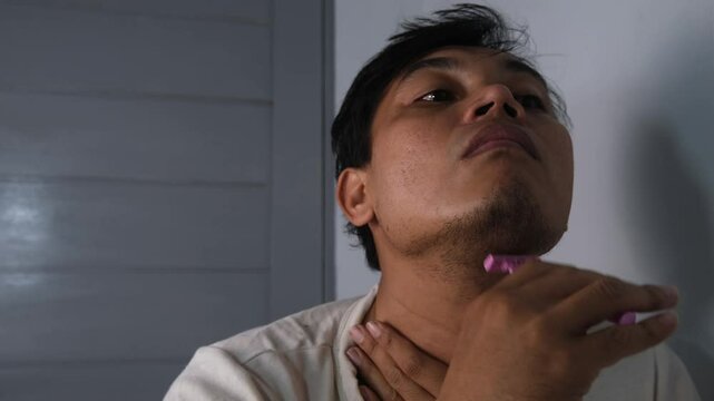 A disheveled Asian man shaving the underside of his beard with a pink manual razor. The scene captures a raw and realistic moment of personal grooming, focusing on the contrast between his rugged appe