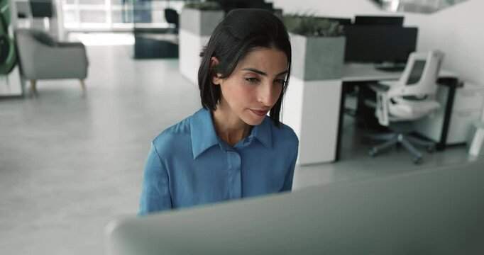 Focused young Latin business professional woman working at desktop in company office alone, looking at monitor, reading on watching online content carefully, testing product