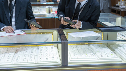 Two jewelry store employees in formal suits checking ring inventory with a clipboard and tablet at a well-lit display counter filled with elegant rings.