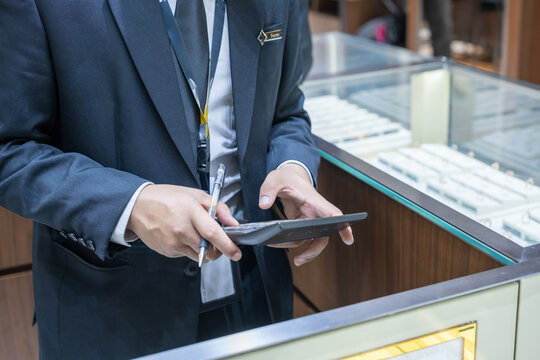 Male staff in suit using calculator and pen for jewelry inventory check at display counter. Business audit, retail management, and product stock control in luxury store environment.