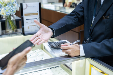Jewelry store transaction scene showing a sales person in a suit holding a calculator and pen while assisting a customer using a smartphone. Modern retail service and luxury shopping moment.


