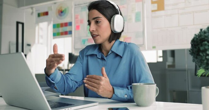Positive beautiful Hispanic business professional woman in headphones speaking on online video conference meeting, using laptop computer at office workplace for Internet communication with partners