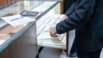 Jewelry store employee organizing gold necklaces in a display case with calculator and inventory sheet, showing retail operations and luxury sales environment.