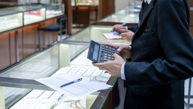 Jewelry store staff in formal attire calculating data with a large calculator and paper sheet near a display case, showing business operations and financial analysis in retail.