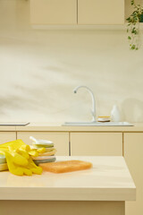 Yellow rubber gloves and cleaning supplies near the sink in a cozy beige kitchen, lifestyle in...