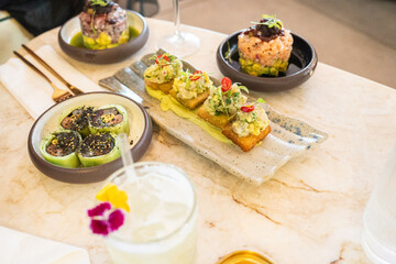 Deliciously arranged small plates featuring fresh seafood, vibrant vegetables, and creative garnishes are enjoyed at a modern bistro, accompanied by a floral cocktail served in a clear glass.