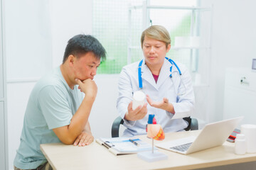 Asian male doctor specializing in cardiology explains heart disease to a patient using a heart model. He provides expert advice, discusses symptoms, risk factors, and treatment in a modern clinic.