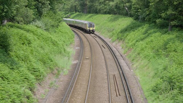 A modern British commuter train departs through green countryside on a curved double-track railway, surrounded by summer foliage and forest under an overcast sky.