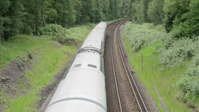 A modern British commuter train departs through green countryside on a curved double-track railway, surrounded by summer foliage and forest under an overcast sky.