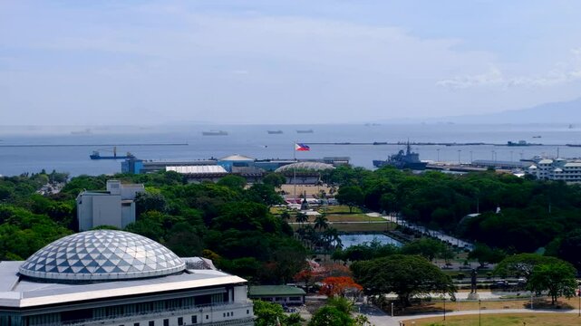 Aerial landscape of Rizal Luneta Park with green trees and flying Philippines flag with view of ocean in capital city Manila
