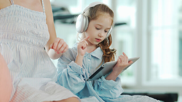 A young girl wearing headphones is engaged with a digital tablet while sitting next to her mother in a bright indoor environment.